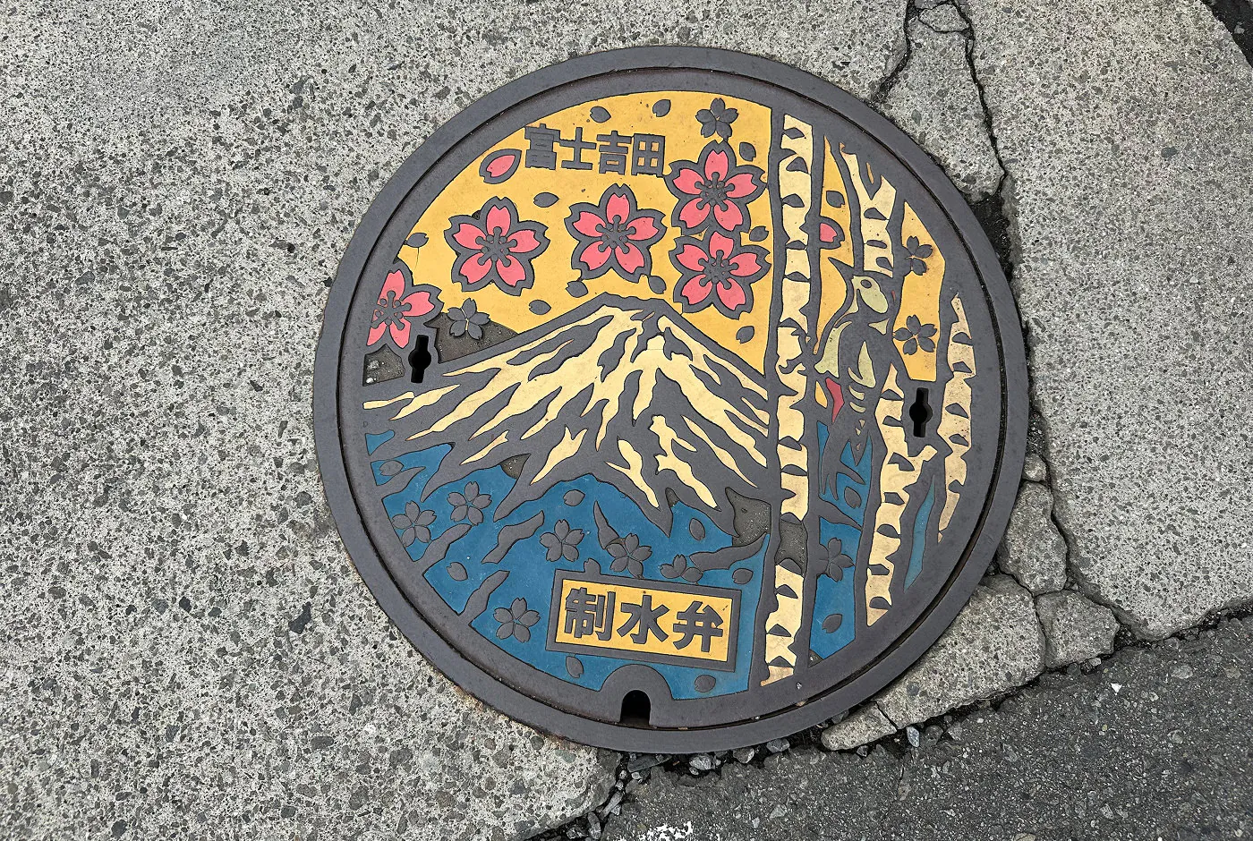 Brunnslock som föreställer vulkanen Mount Fuji. Det har färgerna blått, vitt, gult, rosa och svart.