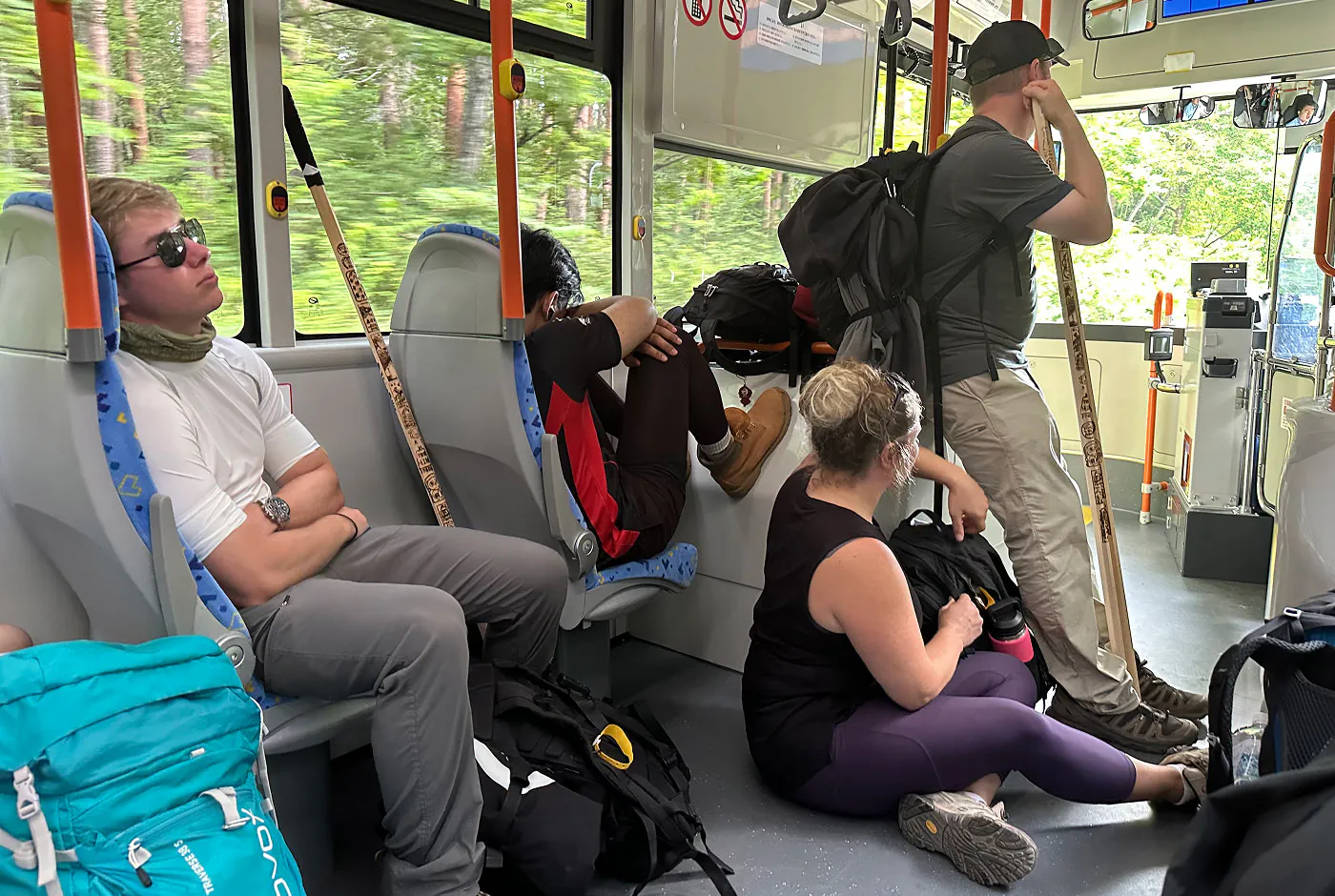 Fyra trötta personer i en buss. Två sitter på säten och sover. En sitter på golvet. En stor lutad mot ett handräcke.
