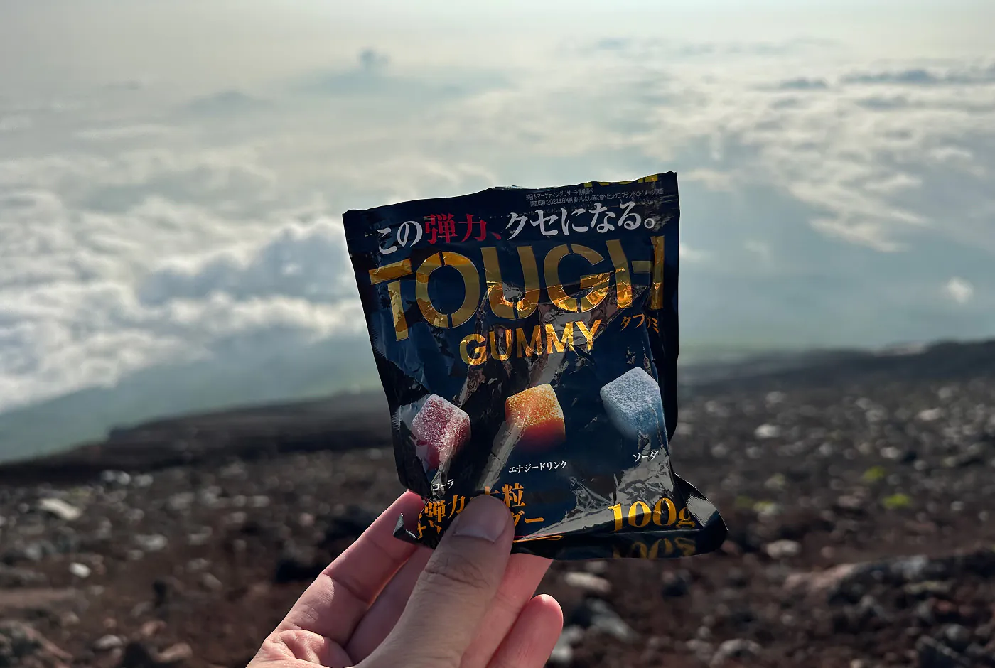 Hand håller i en svart påse med gul text som lyder "Tough Gummy". I bakgrunden syns moln och en bergssluttning.