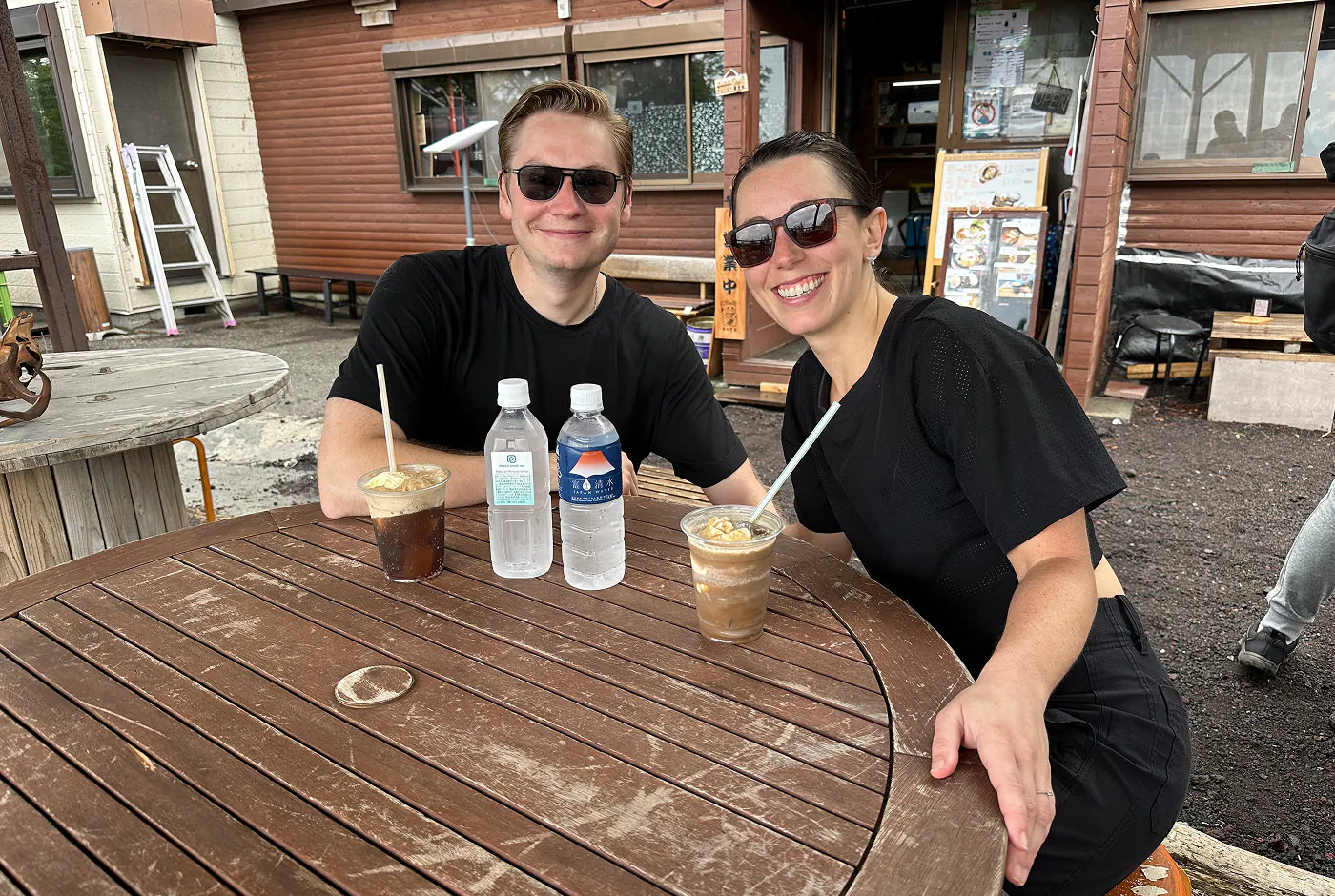 Ungt par med svarta glasögon och svarta kläder dricker varsin läsk med en glassboll i sittande utomhus vid ett träbord.