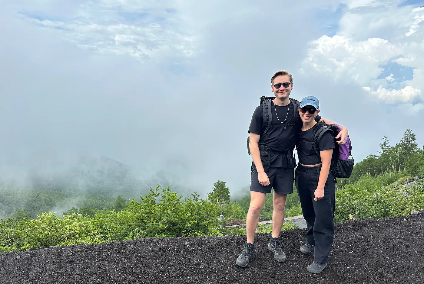 Ungt par med svarta glasögon och svarta kläder håller om varandra på en vandringsled. I bakgrunden syns lite skog i en tjock dimma.