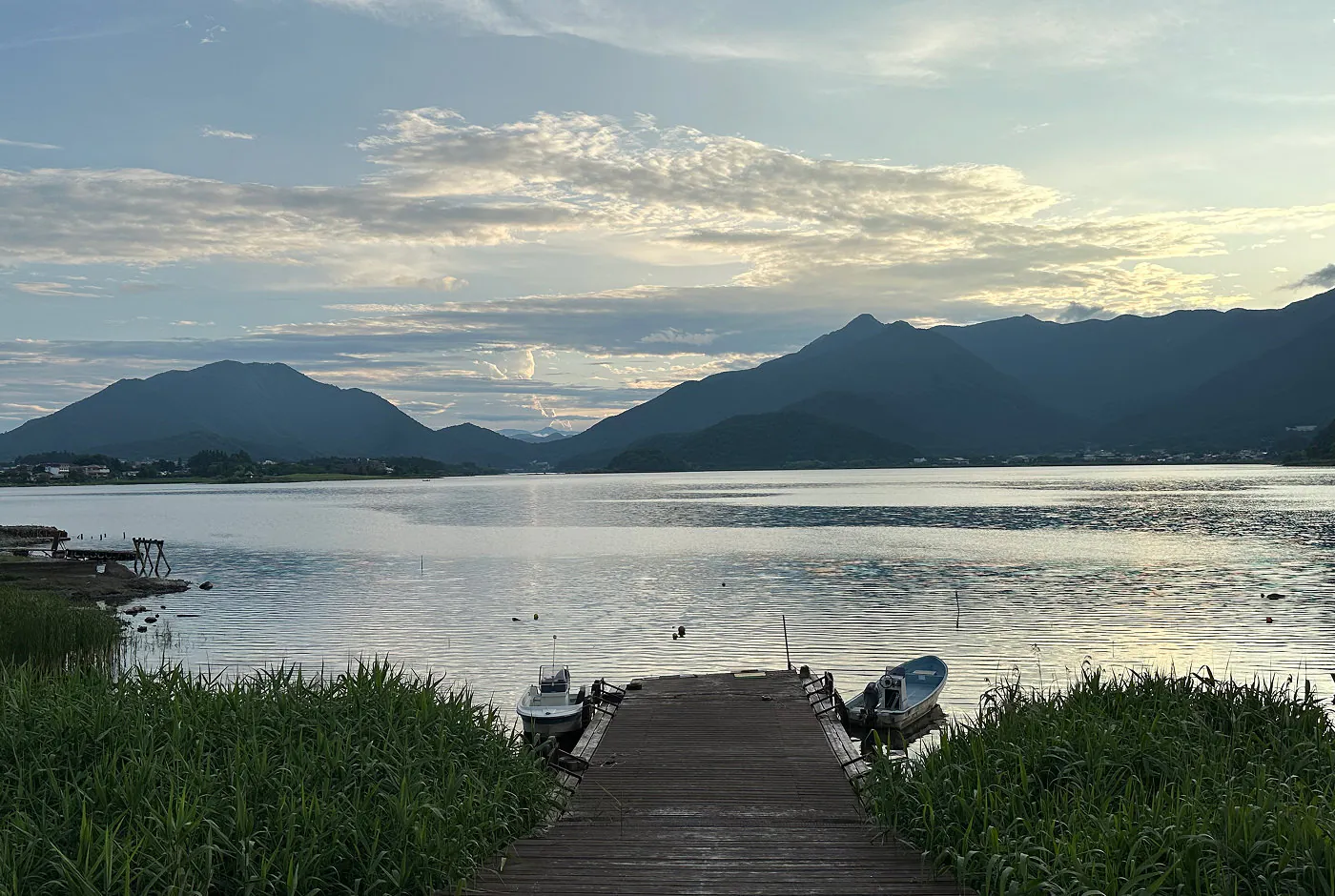 Brygga med två motorbåtar sträcker sig ut på en enorm insjö omgiven av flera lummiga bergskedjor.