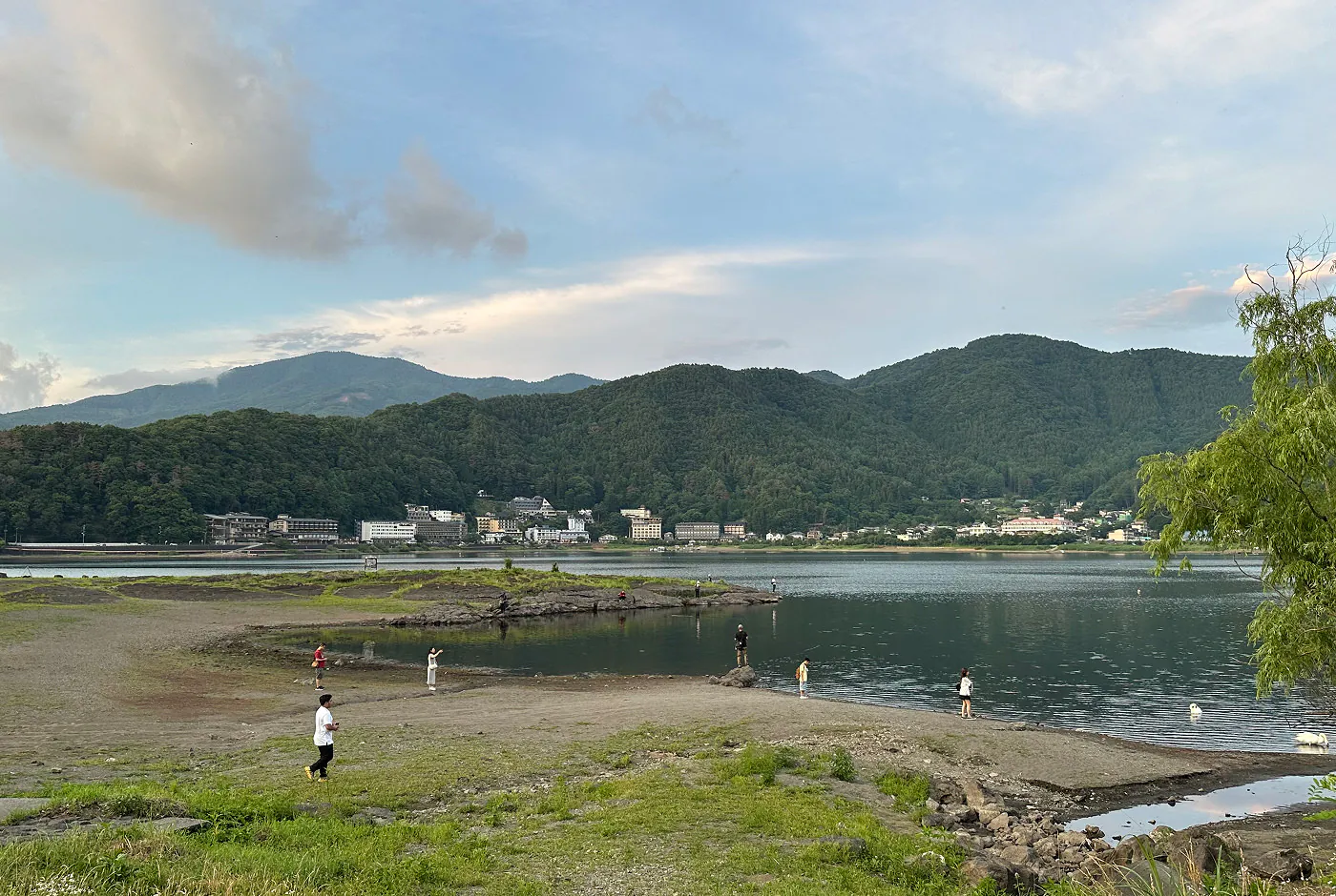 Strandområde vid en stor insjö omgiven av lummiga berg och hus nere vid vattnet. Ett tiotal personer står nere på stranden.