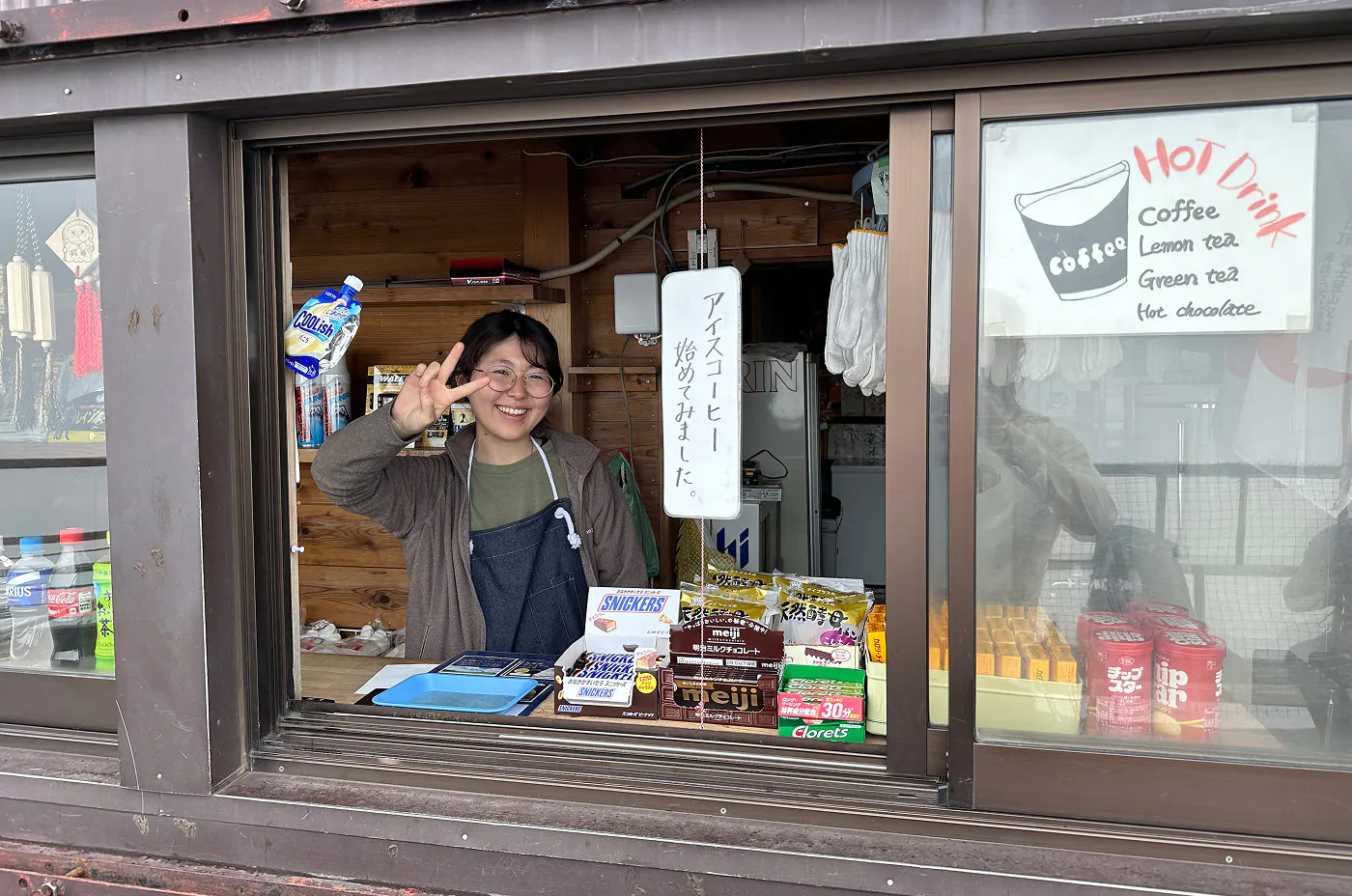 Ung japansk tjej står i en kiosklucka, ler och visar fredstecknet. I lockan finns även en del godis.