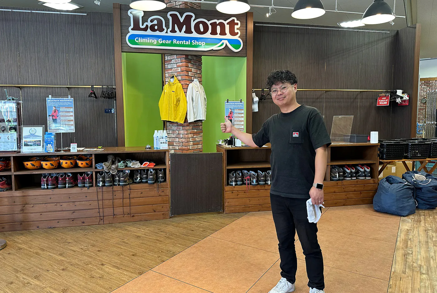 Japansk svartklädd man med glasögon ger tummen upp och ler för kameran. Han står inne i en butik som hyr ut vandringskläder.