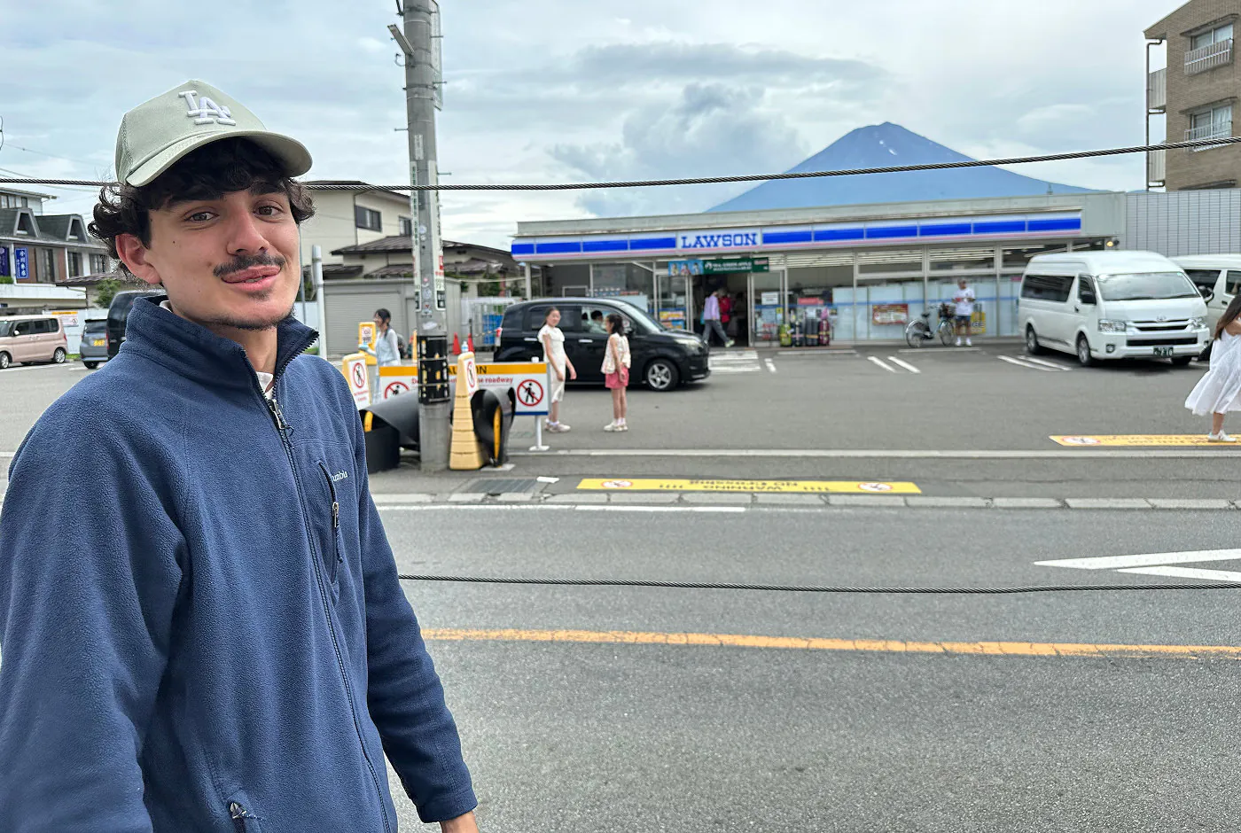 Ung kille med blå tröja och ljus keps ler in i kameran framför en liten matbutik framför Mount Fuji.