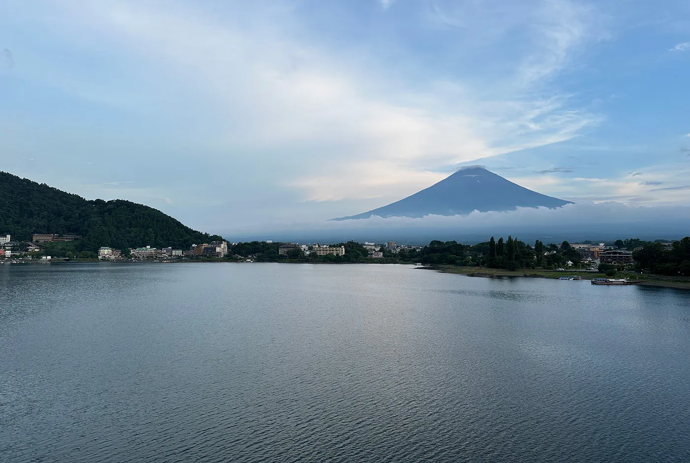 Stor insjö med byggnader längs med vattenbrynet. I fjärran syns enorma Mount Fuji. Moln ligger runt vulkanens fot.
