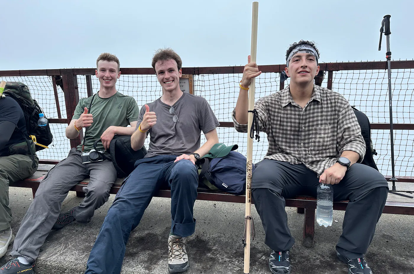 Tre unga killar sitter på en bänk. Två ler och ger tummen upp. En Håller i en vandringspinne och en flaska vatten.