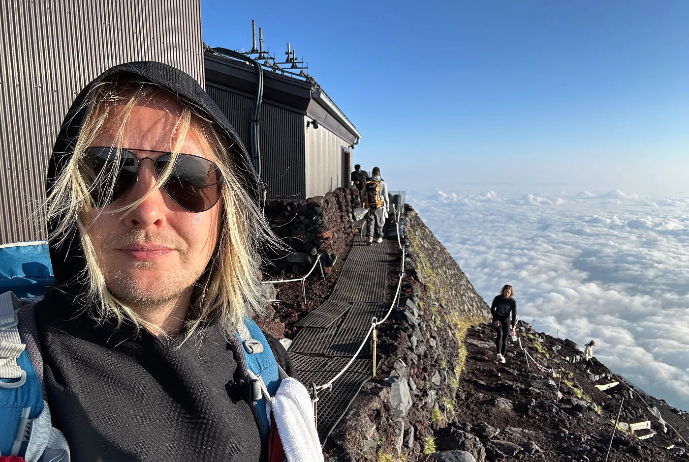 Jag tittar in i kameran vid en hus vid en vandringsled. Jag har svarta solglasögon och en svart uppdragen tröja med huva. I bakgrunden syns moln så långt ögat kan nå.