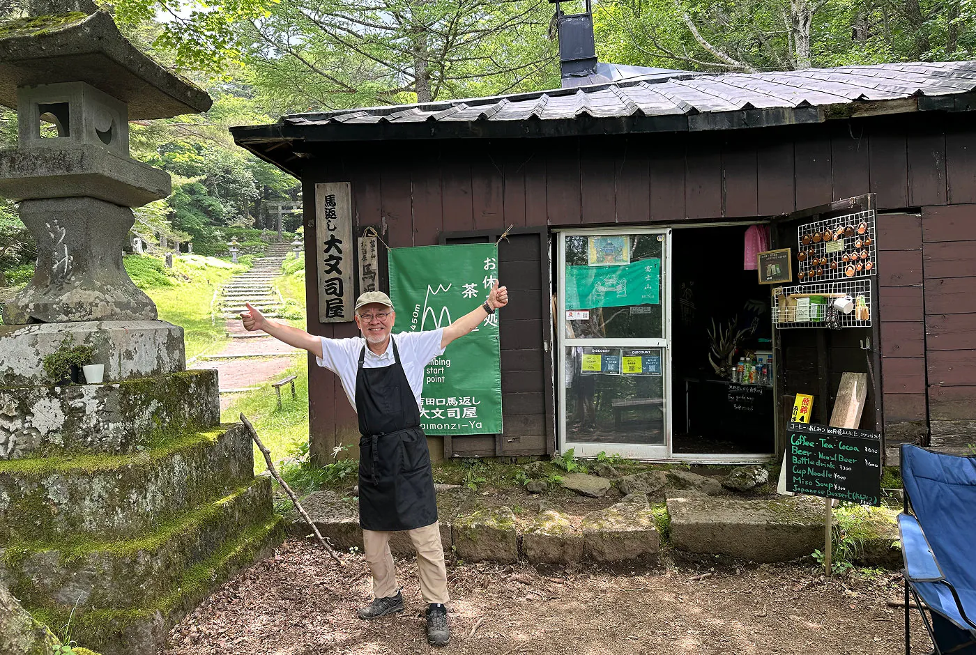 Äldre japansk man med svart förkläde står vid en stuga i en skog och ler samtidigt som han håller upp båda tummarna.