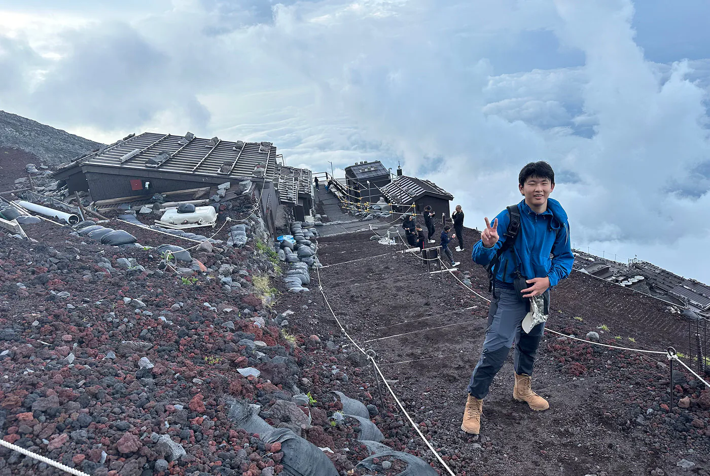 Ung kinesisk kille med blå jacka och mörka byxor visar fredstecknet i början av en stenig vandringsled. I bakgrunden finns några byggnader