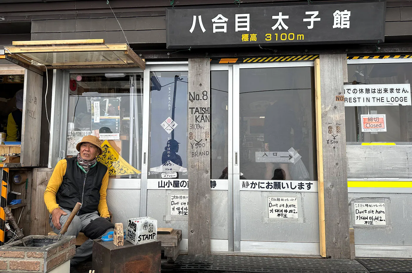 Äldre japansk med med gul mössa och tröja sitter utanför ingången till ett vandrarhem. Ovanför dörren står det "3100 m".