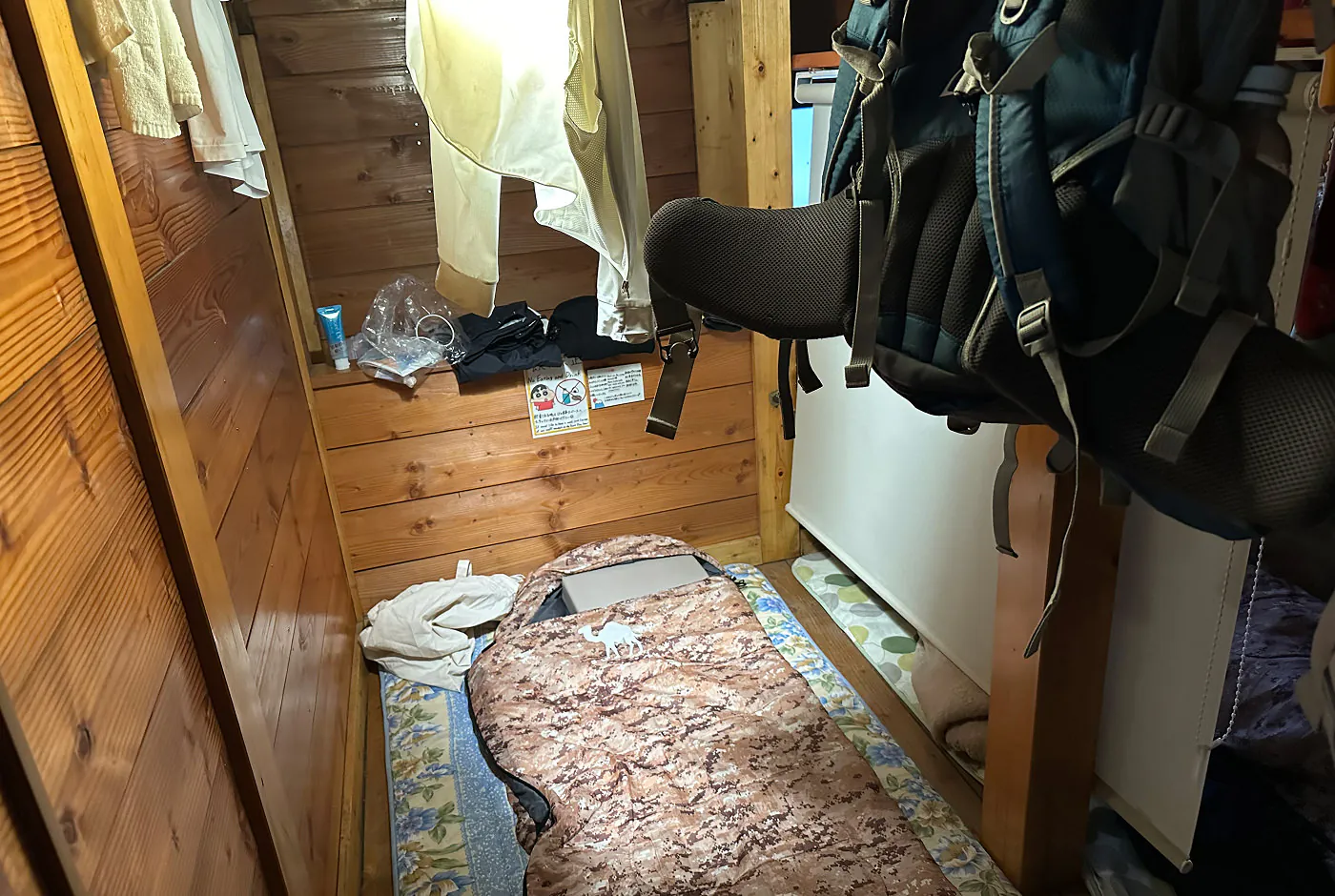 Liten sovplats i ett vandrarhem. På golvet ligger en sovsäck och på en hylla ovanför den ligger lite kläder. I förgrunden hänger en ryggsäck från taket.