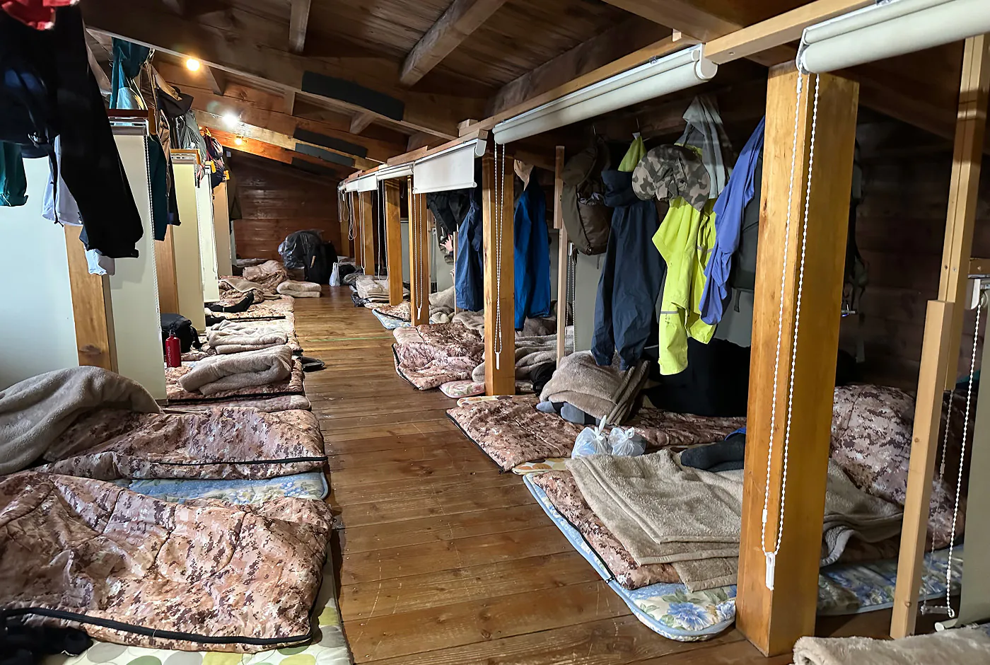 Lång sovsal i en stuga. På golvet ligger sovsäckar jämte varandra och kläder hänger från taket. Det ser mysigt ut.