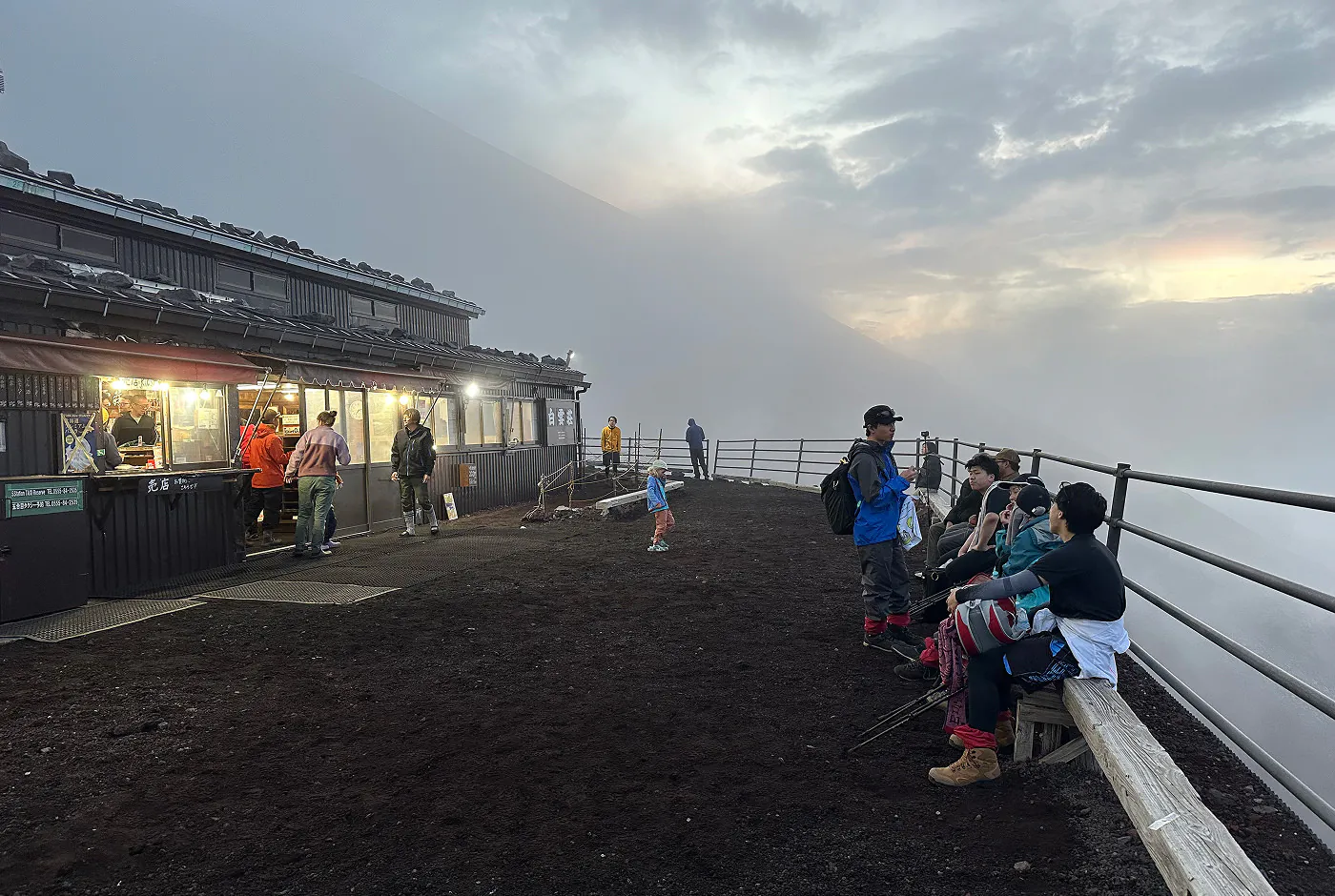 Grusterass utanför ett vandrarhem på ett berg. Folk står ute vid terassens räck och inne i stugan. Himlen är molnig och det börjar bli mörkt.