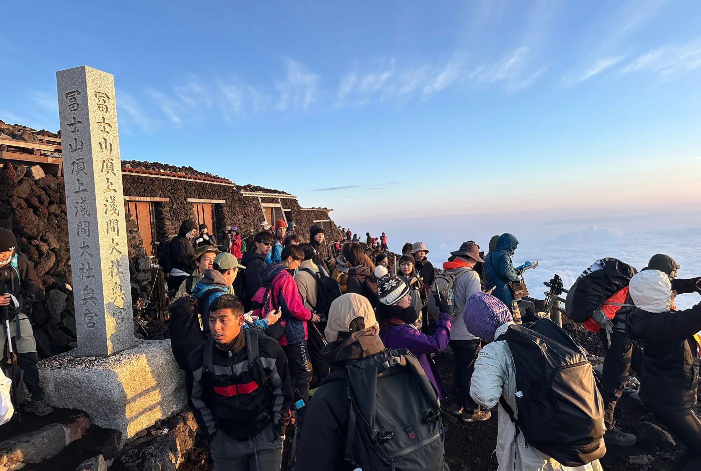 Folkmassa på toppen av Mount Fuji. Alla har färggranna träningskläder på sig. Himlen är klarblå.