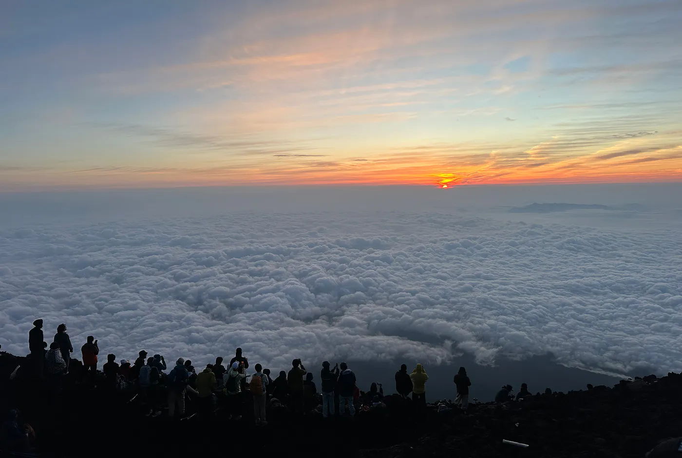 Ett femtiotal personer står på ändan av en bergstopp och tittar på ett molnlandskap. I fjärran går solen upp.