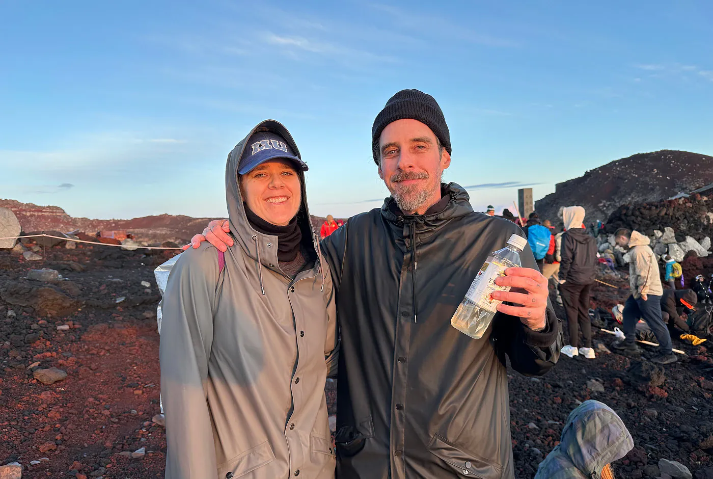 En man och en kvinna i gråa regnkläder och mössor håller om varandra och ler in i kameran. En av dem håller i en flaska.