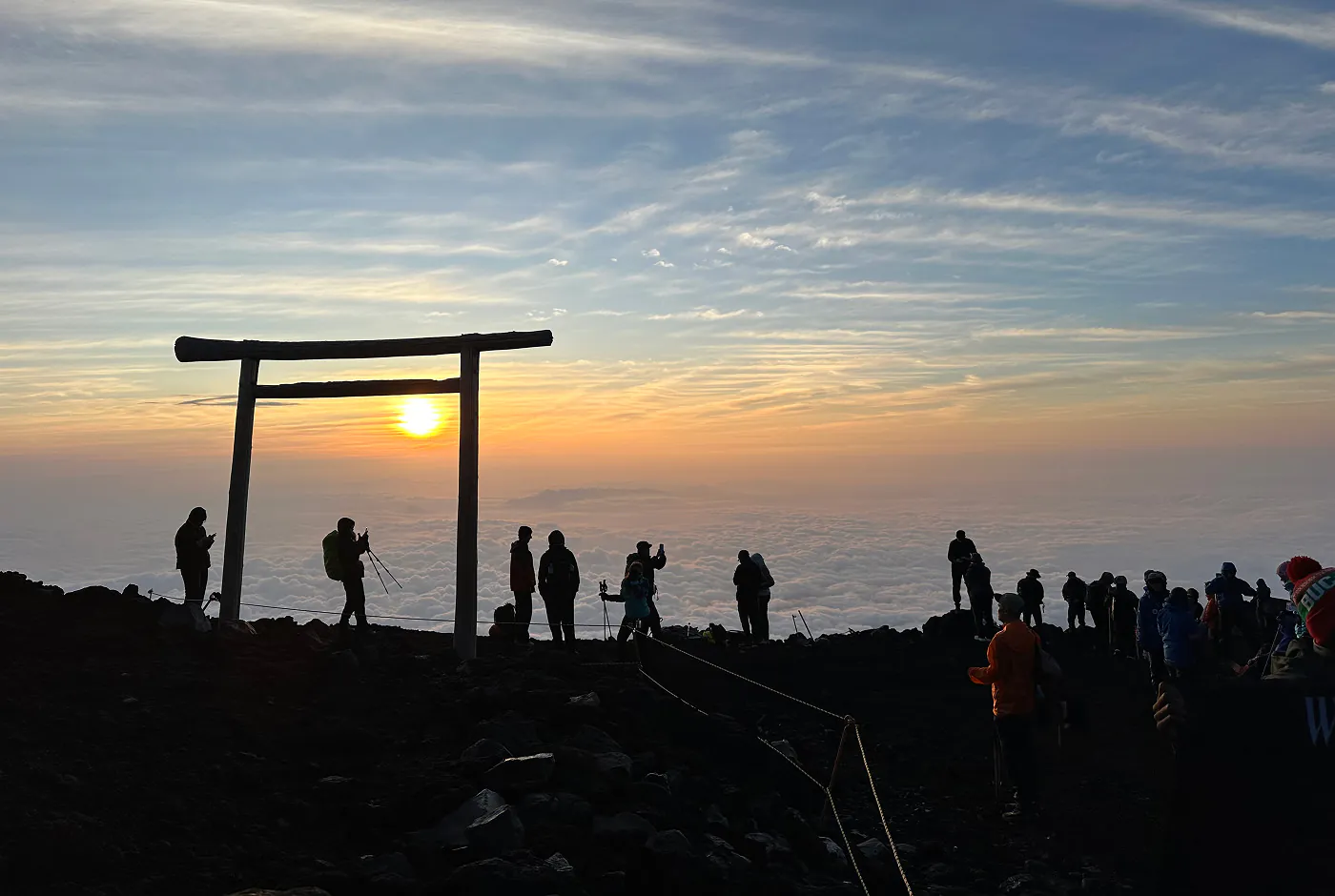 Det är tidig morgon och solen går upp över Mount Fuji. I förgrunden ser man siluetten av många människor och en torii-port.