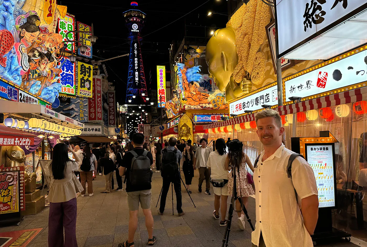 Ung blond kille med mustasch och vit skjorta står på en väldigt upplyst och trångt gata om en stativkamera. I fjärran syns ett upplyst torn.