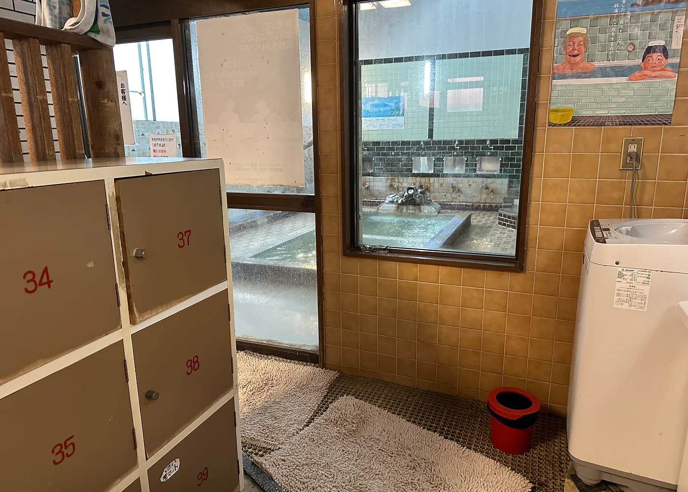 Omklädningsrum med skåp och tvättmaskin i ett litet badhus. Via ett fönster kan man se badet som har en rektangulär pool i mitten.