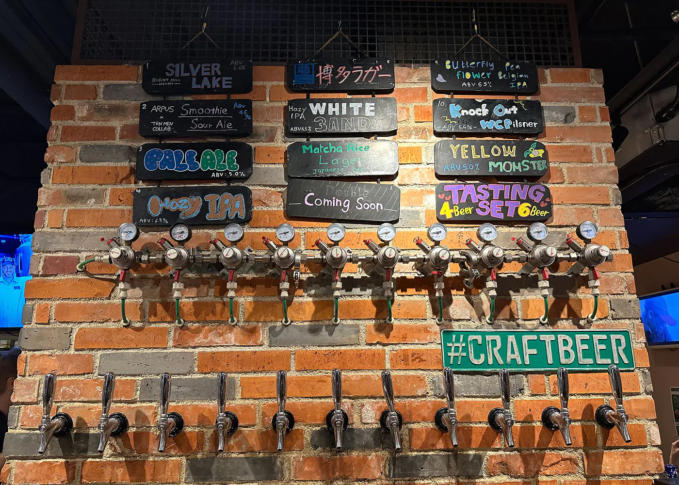 Tegelvägg i en bar. På väggen hänger tolv skyltar med olika ölsorter och under det tappar för dessa sorter.