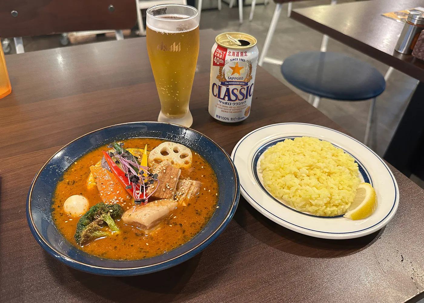 En mörk skål med orange currysoppa med grönsaker står på ett bord bredvid en vit tallrik med ris och ett upphällt ölglas.