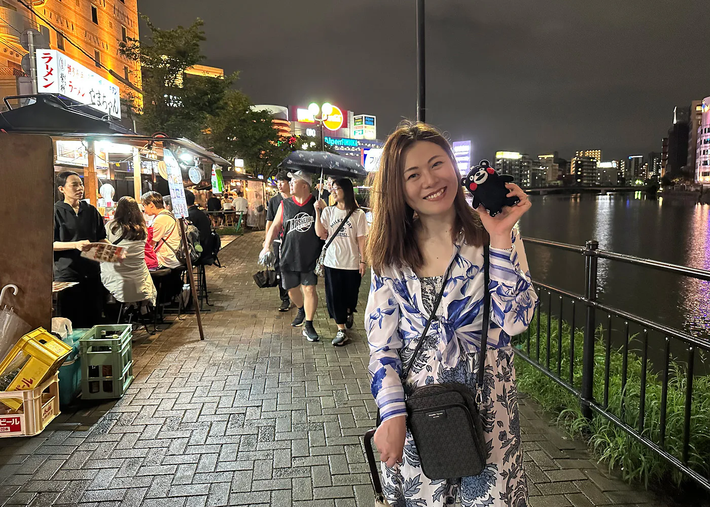 Vacker japansk kvinna i ljus klänning ler in i kameran. Hon står på en gågata med matstånd längd med en flod.