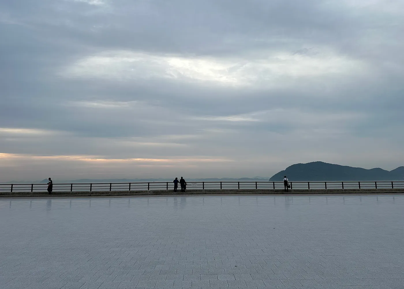 Stor och bred stenpromenad vid ett hav. Fyra personer står vid räcket vid havet. I fjärran skymtar lummiga stora öar.