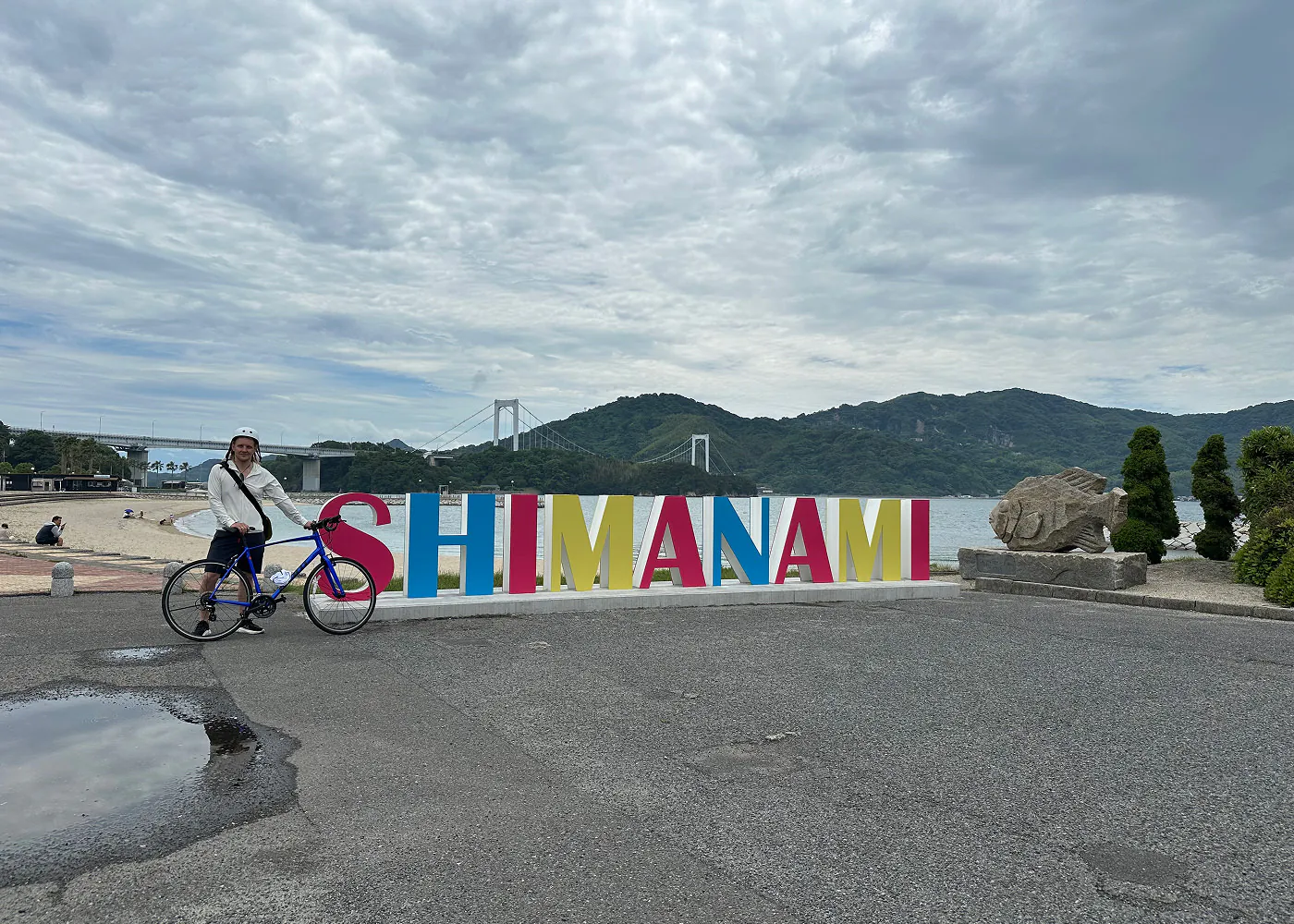 Jag står och håller i en cykel framför en skylt framför en sandstrand. Skylten lyder "Shimanami". I fjärran ser man en hängbro och lummiga berg.
